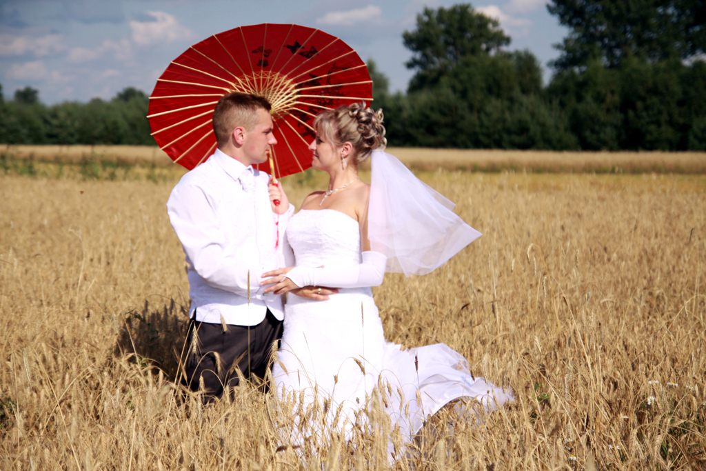 Fotografia pary młodej na polu w zbożu z czerwoną parasolką autorstwa Ani Wilanowskiej.